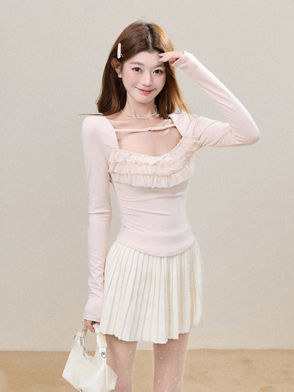 A young woman posing in a light pink long-sleeve top with a ruffled sweetheart neckline and a cream pleated mini skirt, holding a small cream handbag against a neutral beige background.