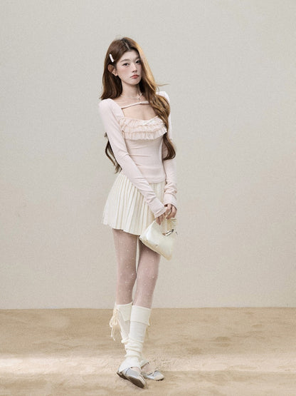 A young woman models a light pink long-sleeve top with a ruffled sweetheart neckline, paired with a matching pleated mini skirt, white thigh-high socks, and a small cream-colored handbag, set against a minimalist beige studio backdrop.