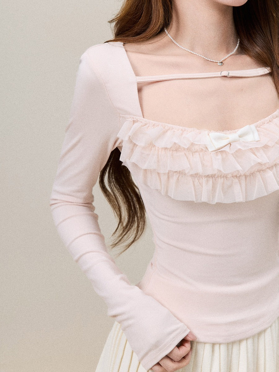 A close-up of a model wearing a pale pink long-sleeve top with a square neckline, ruffled trim, a bow detail, and a thin strap across the chest.