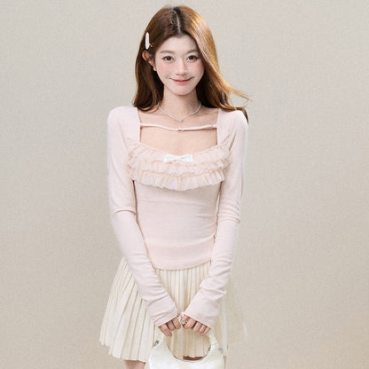 A young woman stands against a plain beige studio wall wearing a pale pink long-sleeve top with a ruffled chest detail, a cream pleated skirt, sheer polka-dot tights, and holding a small cream handbag.