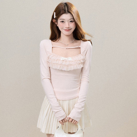 A young woman stands against a plain beige studio wall wearing a pale pink long-sleeve top with a ruffled chest detail, a cream pleated skirt, sheer polka-dot tights, and holding a small cream handbag.