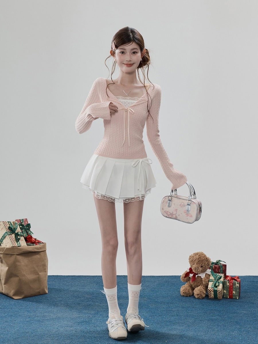 A young woman stands in a studio wearing a light pink ribbed long-sleeve top with a lace-up sweetheart neckline, a white pleated mini skirt with lace trim, white socks, and white sneakers. She holds a small floral-patterned handbag.