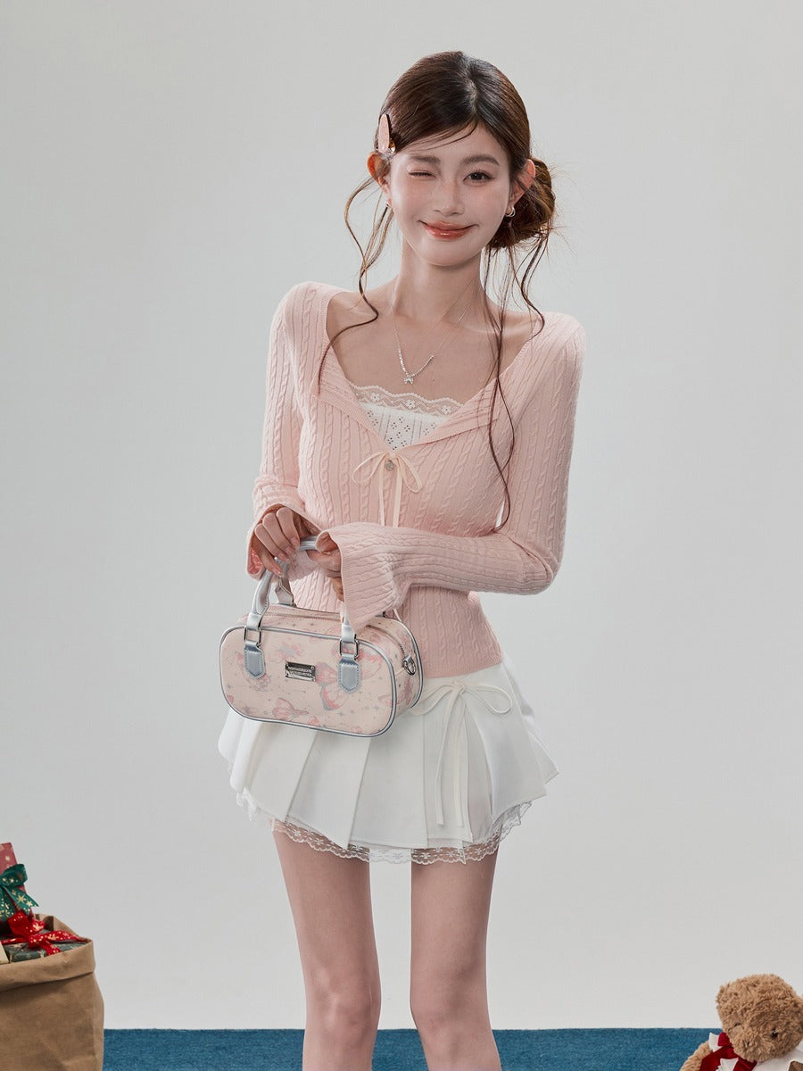 A young woman models a light pink ribbed knit long-sleeve top with a lace-trimmed scoop neckline, paired with a white pleated mini skirt and holding a small pink handbag against a plain light gray studio background.