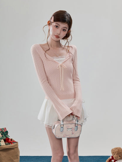 A young woman models a light pink ribbed long-sleeve knit top with a lace-trimmed sweetheart neckline and a front tie detail, paired with a white pleated mini skirt and holding a small pink patterned handbag against a plain light gray studio background.