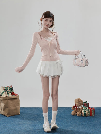 A young woman models a light pink ribbed knit long-sleeve top with a white lace-trimmed mini skirt, white socks, and white sneakers. She holds a small pink floral handbag against a clean studio backdrop with holiday props on the floor.