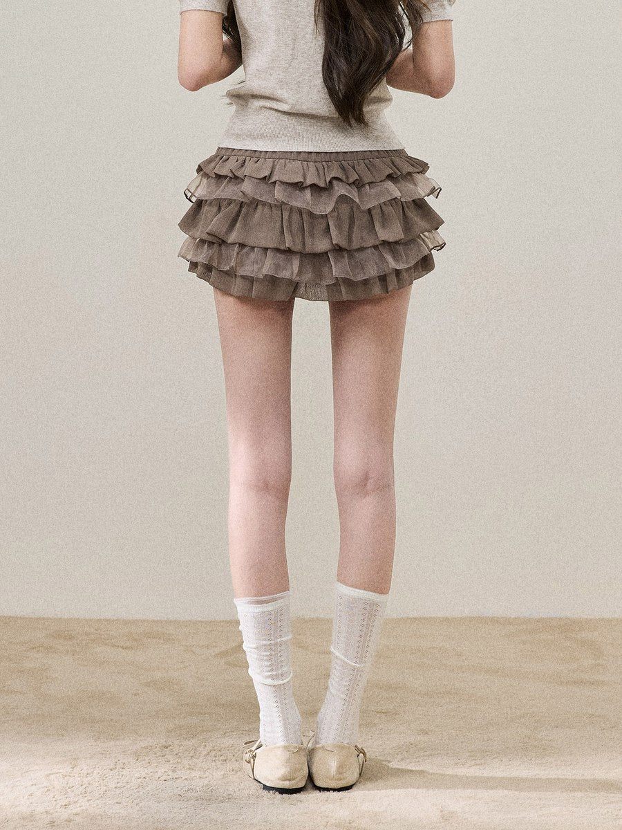 A model from the back wearing a brown ruffled tiered mini skirt, a light beige short-sleeve top, white socks, and beige shoes, standing against a neutral beige background.