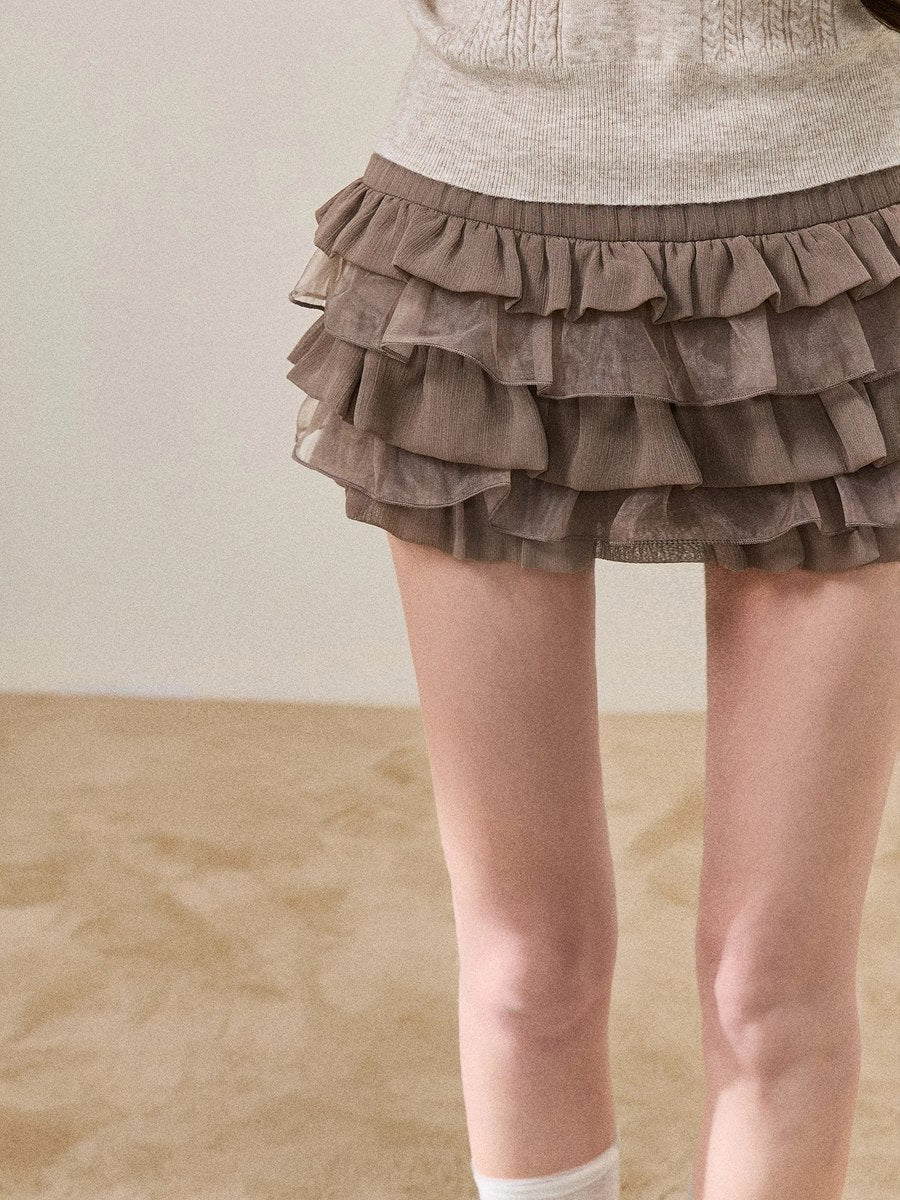 A close-up of a person wearing a tiered ruffle mini skirt in taupe brown, paired with a light beige knit top and white socks.