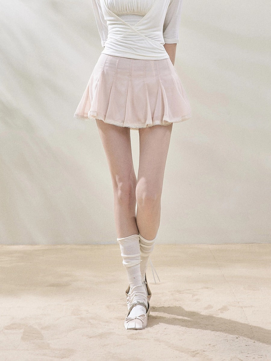 A model from the waist down wearing a light pink pleated mesh mini skirt, a white ribbed top, white knee-high socks, and white lace-up dance shoes, standing against a neutral beige backdrop.