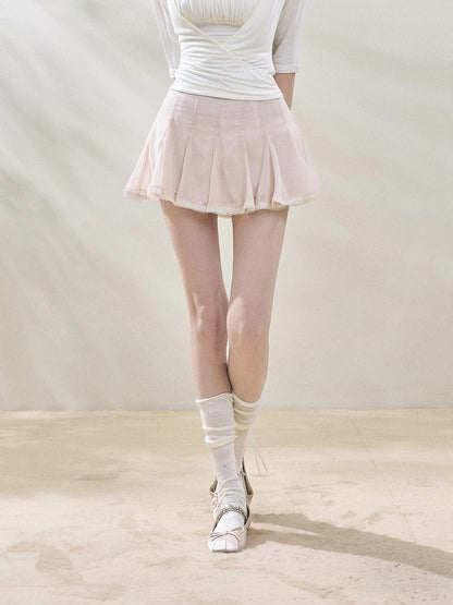 A model from the waist down wearing a light pink pleated mesh mini skirt, a white ribbed top, white knee-high socks, and white lace-up dance shoes, standing against a neutral beige backdrop.