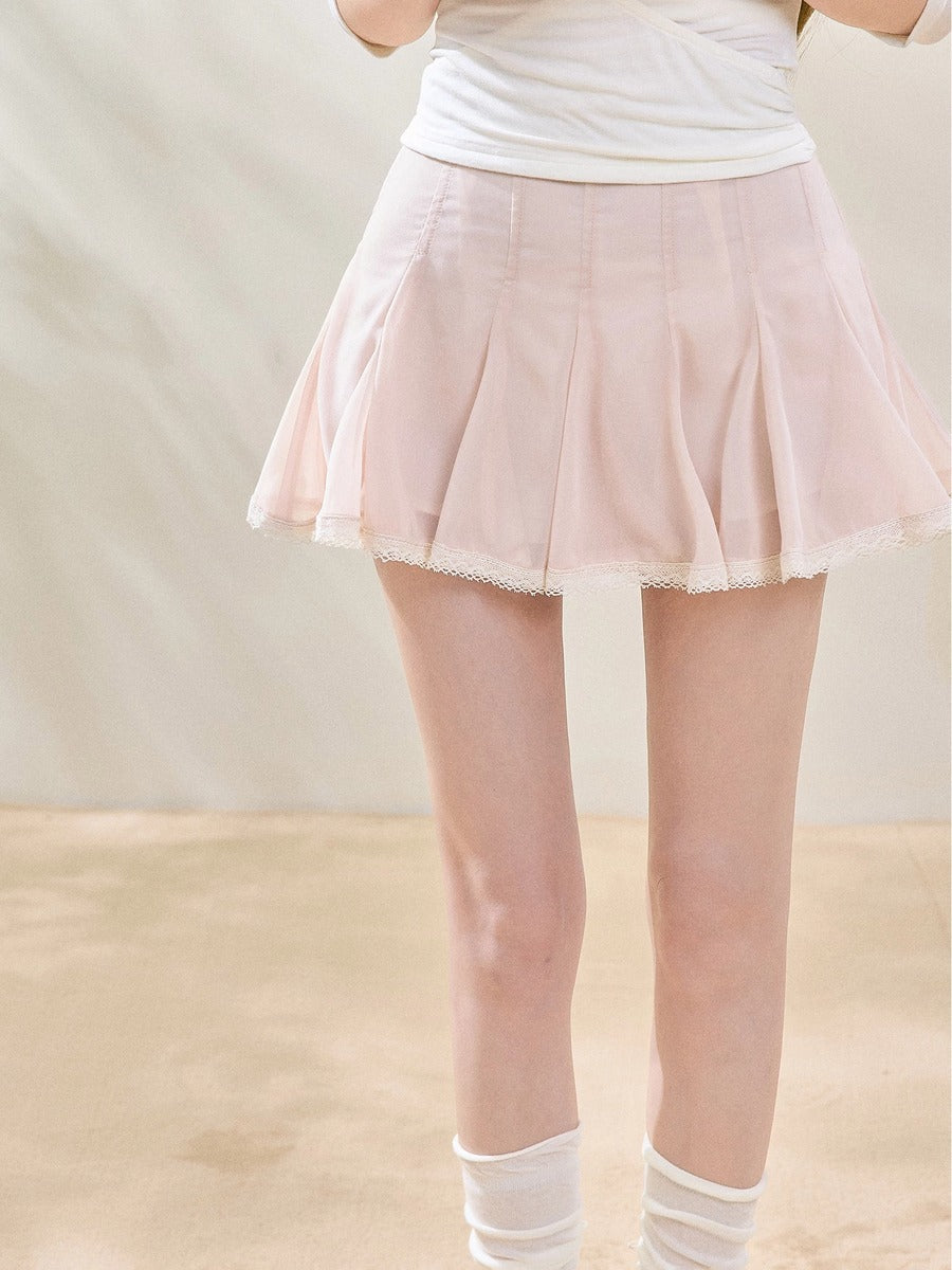 A light pink pleated mini skirt with a white lace hem, modeled with a white top and white socks against a neutral beige background.