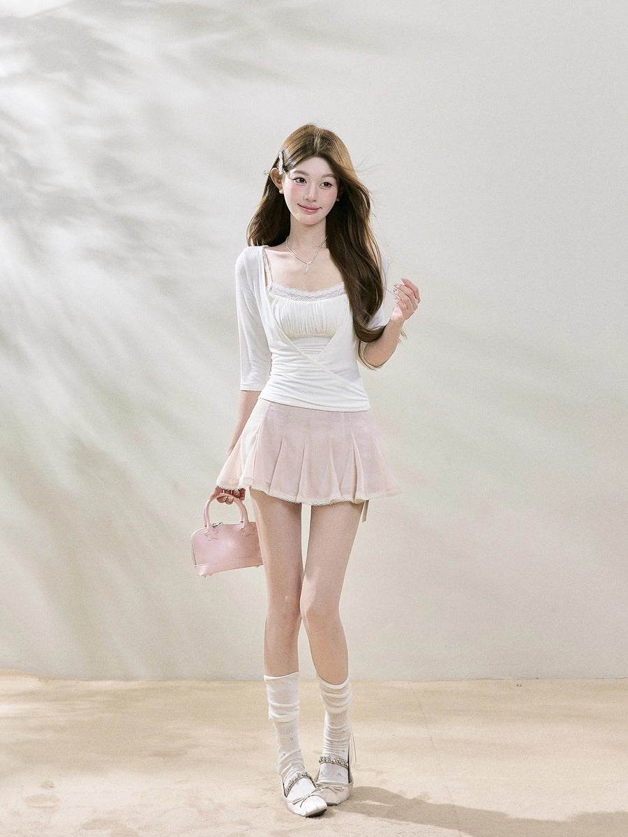 A young woman stands in a studio wearing a white ribbed long-sleeve top and a light pink pleated mini skirt, holding a small pink handbag. She is wearing white knee-high socks and white sneakers.
