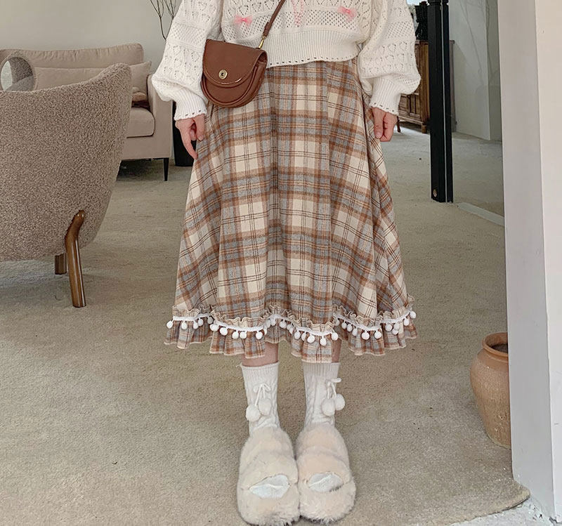 Person wearing a plaid skirt with pom-pom trim, white sweater, and slippers indoors.