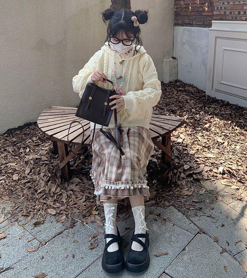 Child in a cream sweater and plaid skirt holding a black handbag on a leaf-covered ground.