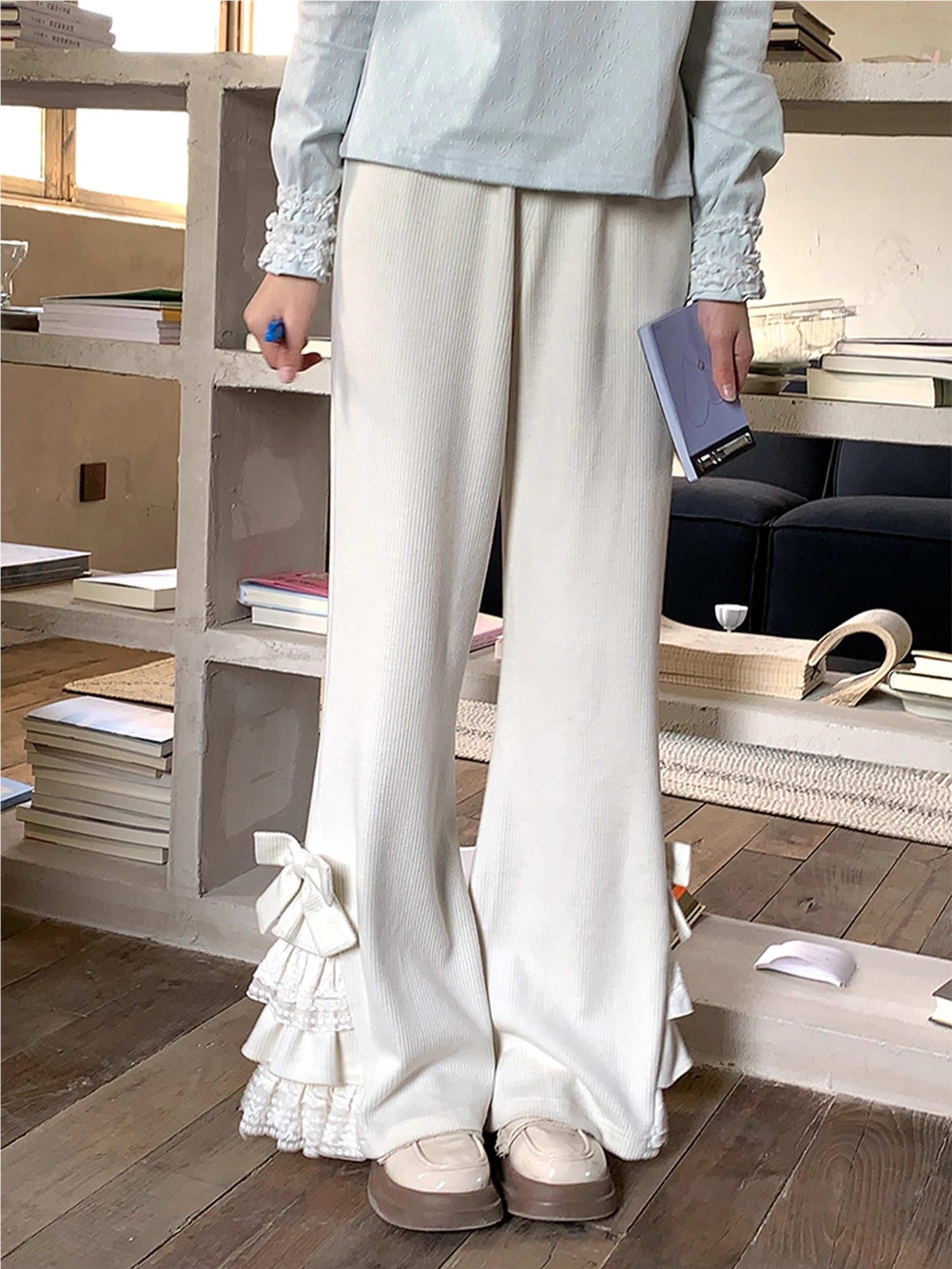 Person wearing light-colored pants with ruffled details in a room with wooden flooring and shelves.