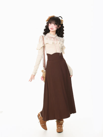 A young woman models a cute, retro-inspired outfit featuring a cream lace long-sleeve top with ruffles and a high-waisted brown pleated maxi skirt. She is styled with brown suede boots, a small brown crossbody bag, and a brown hair accessory, set against a seamless white studio background.