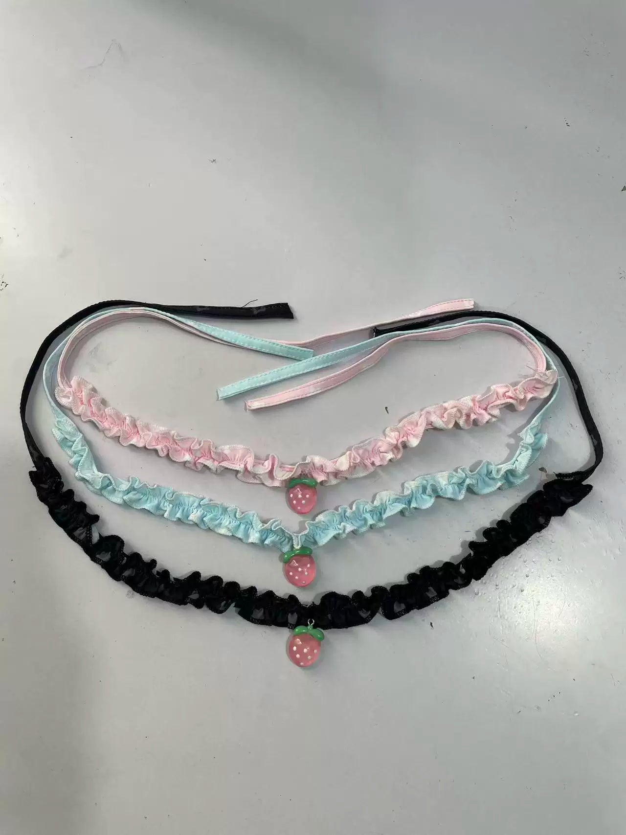 Three ruffled garters laid flat on a light gray surface: one black, one light pink, and one light blue, each with a small strawberry charm at the center.