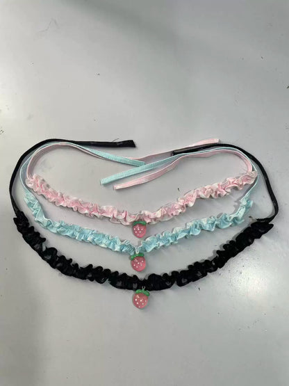 Three ruffled garters laid flat on a light gray surface: one black, one light pink, and one light blue, each with a small strawberry charm at the center.