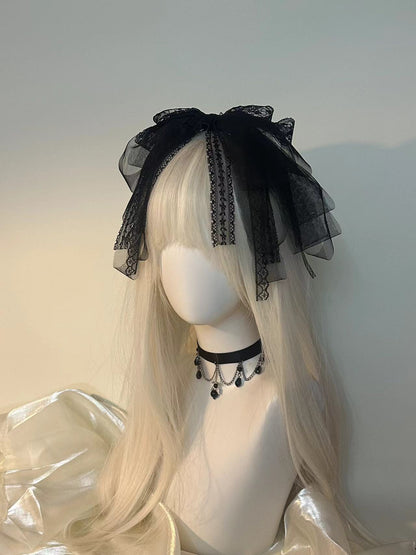 A long platinum blonde wig with straight bangs displayed on a white mannequin head, styled with a large black sheer tulle bow headpiece and a black choker necklace with dangling black beads.