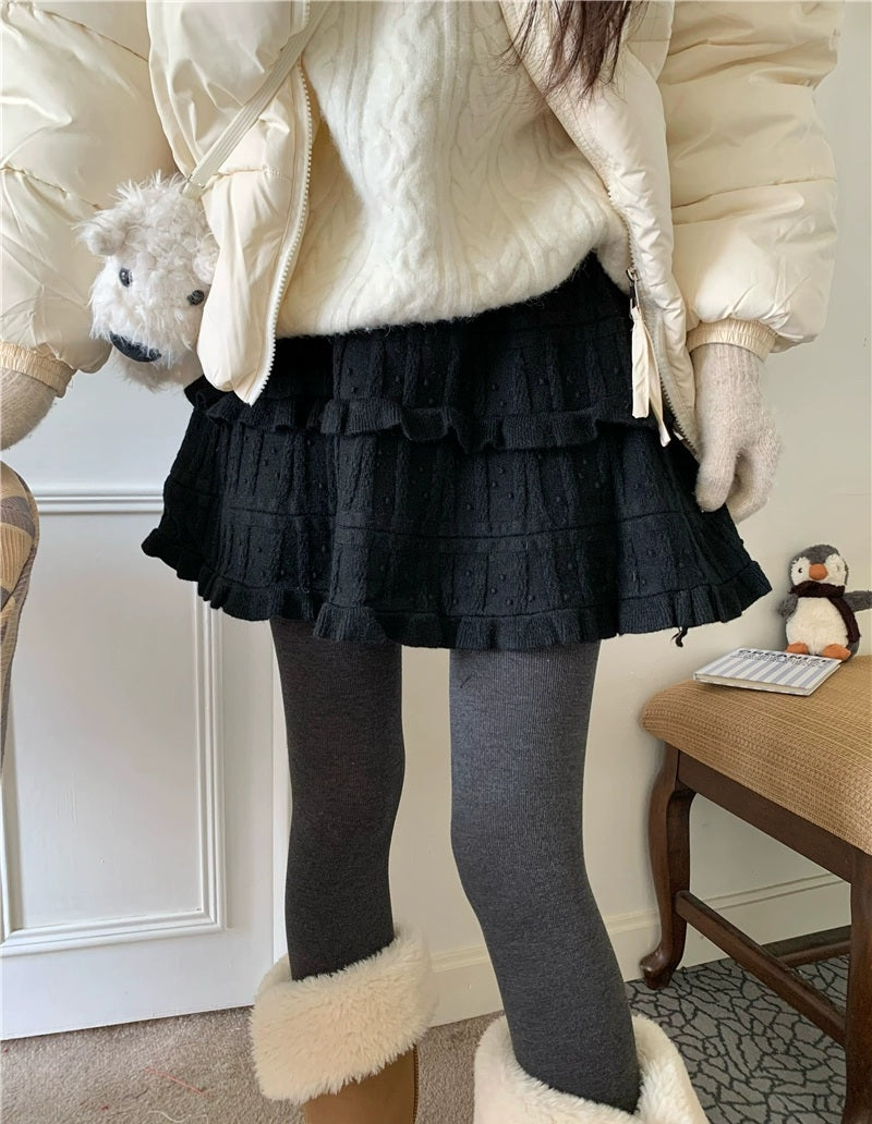 Person wearing a white coat with fur-lined cuffs, black ruffled skirt, and gray tights in an indoor setting.
