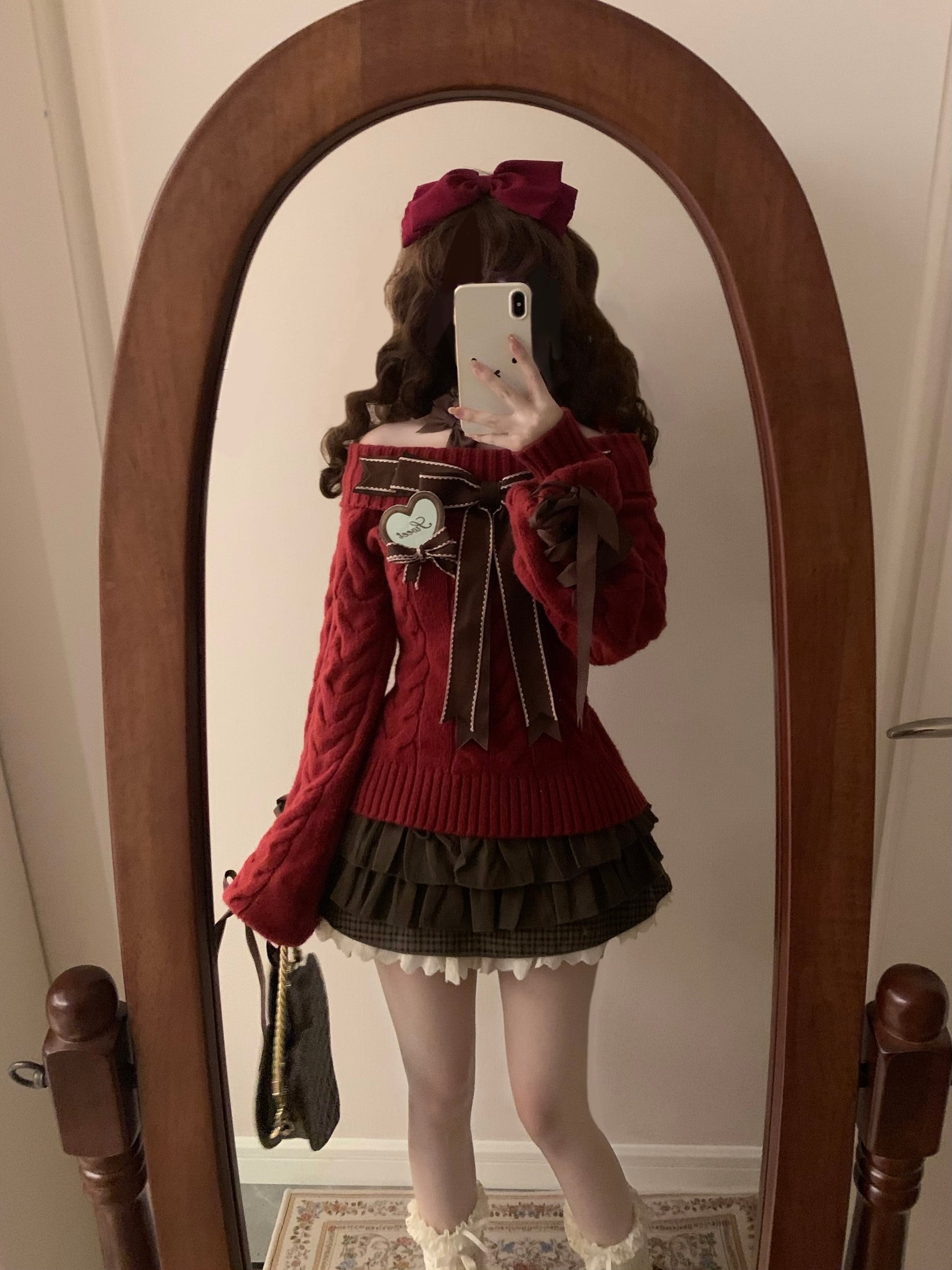Person wearing a red sweater with large bows and a dark skirt, standing in front of a mirror.