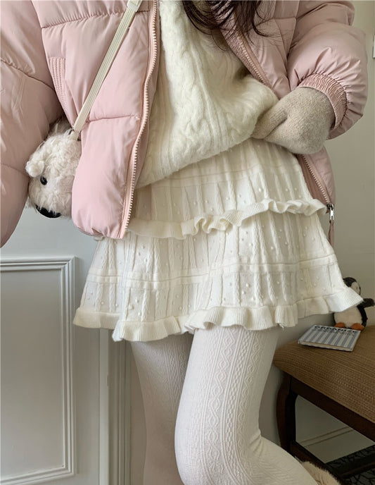 Person wearing a pink puffer jacket, white sweater, and cream-colored skirt holding a teddy bear.