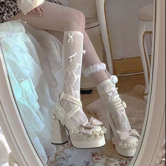 White lace-up high-heeled shoes with decorative elements worn by a person sitting in front of a mirror.