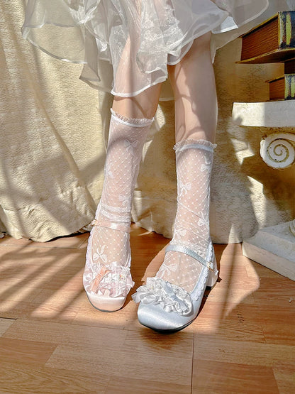 Silver high-heeled shoes with lace details worn with a white dress on a wooden floor.
