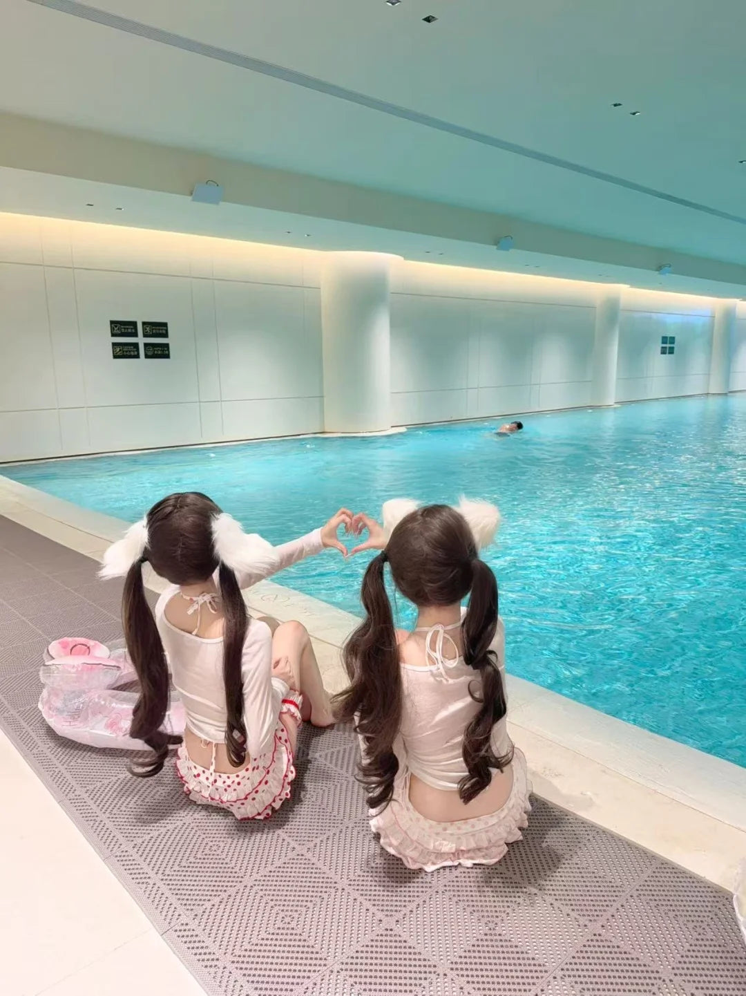 Two young girls with pigtails sitting by an indoor pool, making a heart shape with their hands.