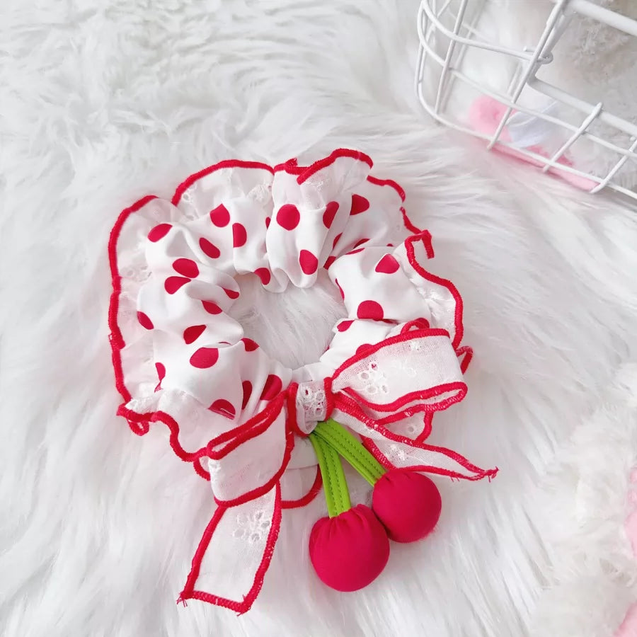 White scrunchie with red polka dots and cherry decorations on a white fluffy surface.