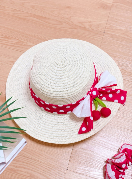 White straw hat with a red polka dot ribbon and cherry decorations on a wooden surface.