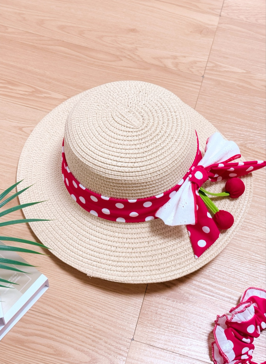 Beige straw hat with a red polka dot ribbon and cherry decorations on a wooden surface.