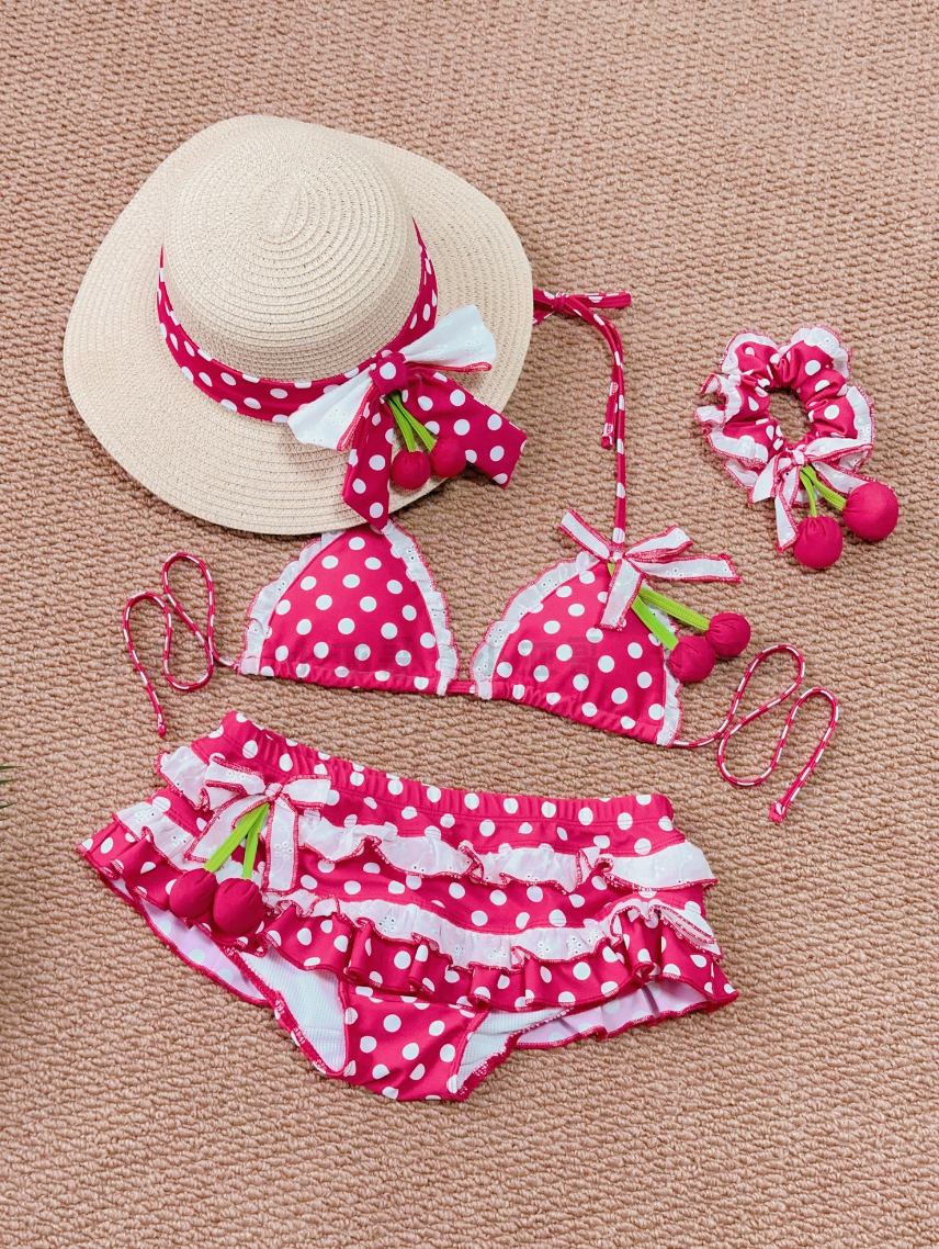 Red polka dot bikini set with cherry details, matching hat, and hair accessory on a beige background.
