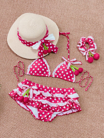 Red polka dot bikini set with cherry details, matching hat, and hair accessory on a beige background.