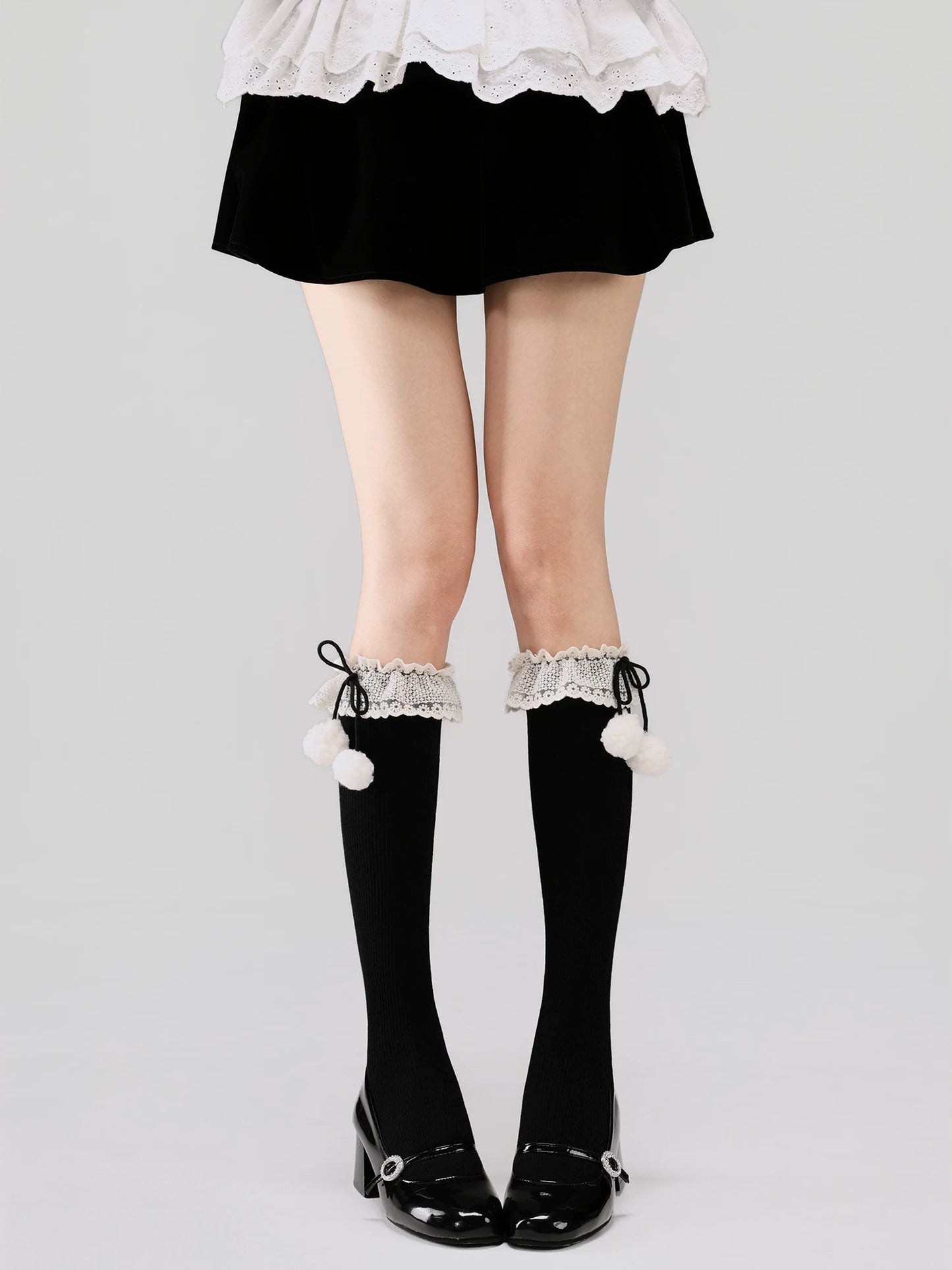 Black knee-high socks with white lace trim and pom-poms worn with a black skirt and white shoes on a light gray background.