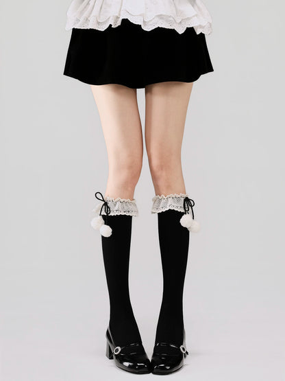Black knee-high socks with white lace trim and pom-poms worn with a black skirt and white shoes on a light gray background.