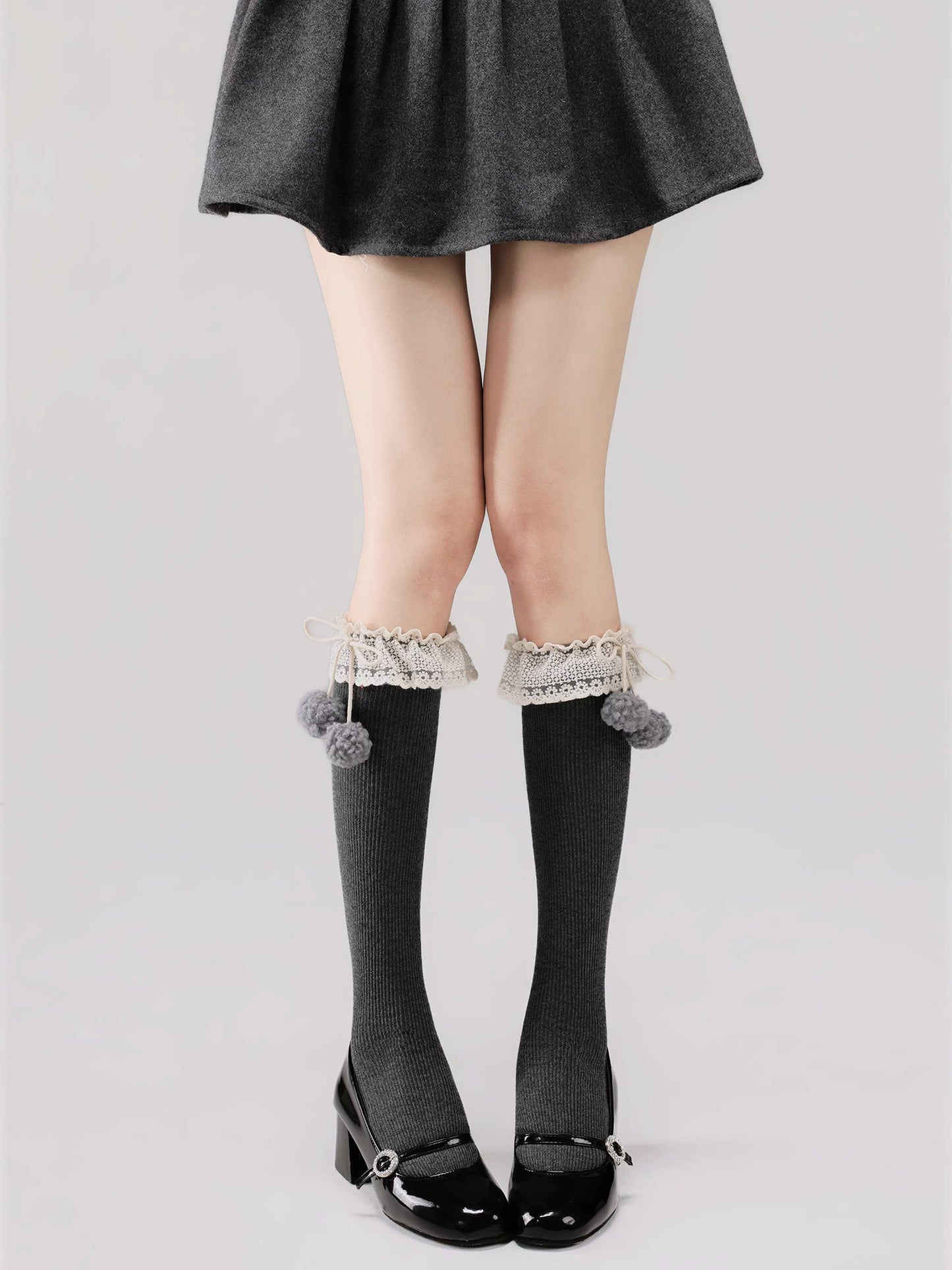 Person wearing gray knee-high socks with lace trim and pom-poms, paired with a black skirt and shoes on a light gray background.