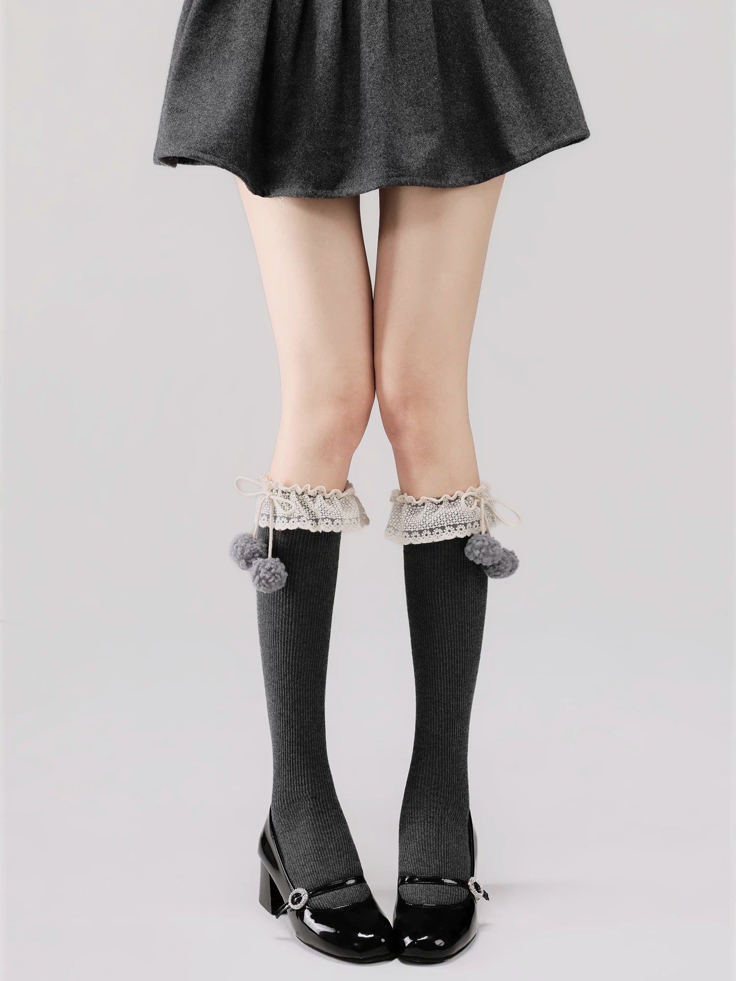 Person wearing gray knee-high socks with lace trim and pom-poms, paired with a black skirt and shoes on a light gray background.