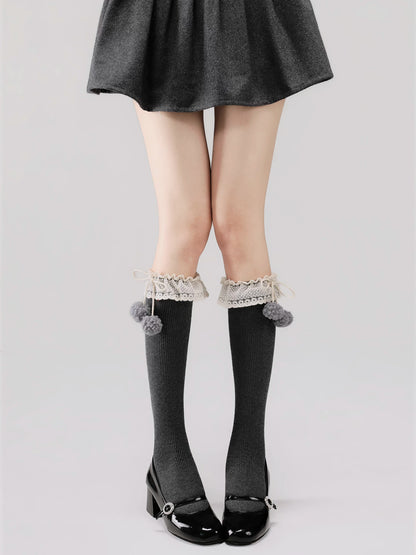Person wearing gray knee-high socks with lace trim and pom-poms, paired with a black skirt and shoes on a light gray background.