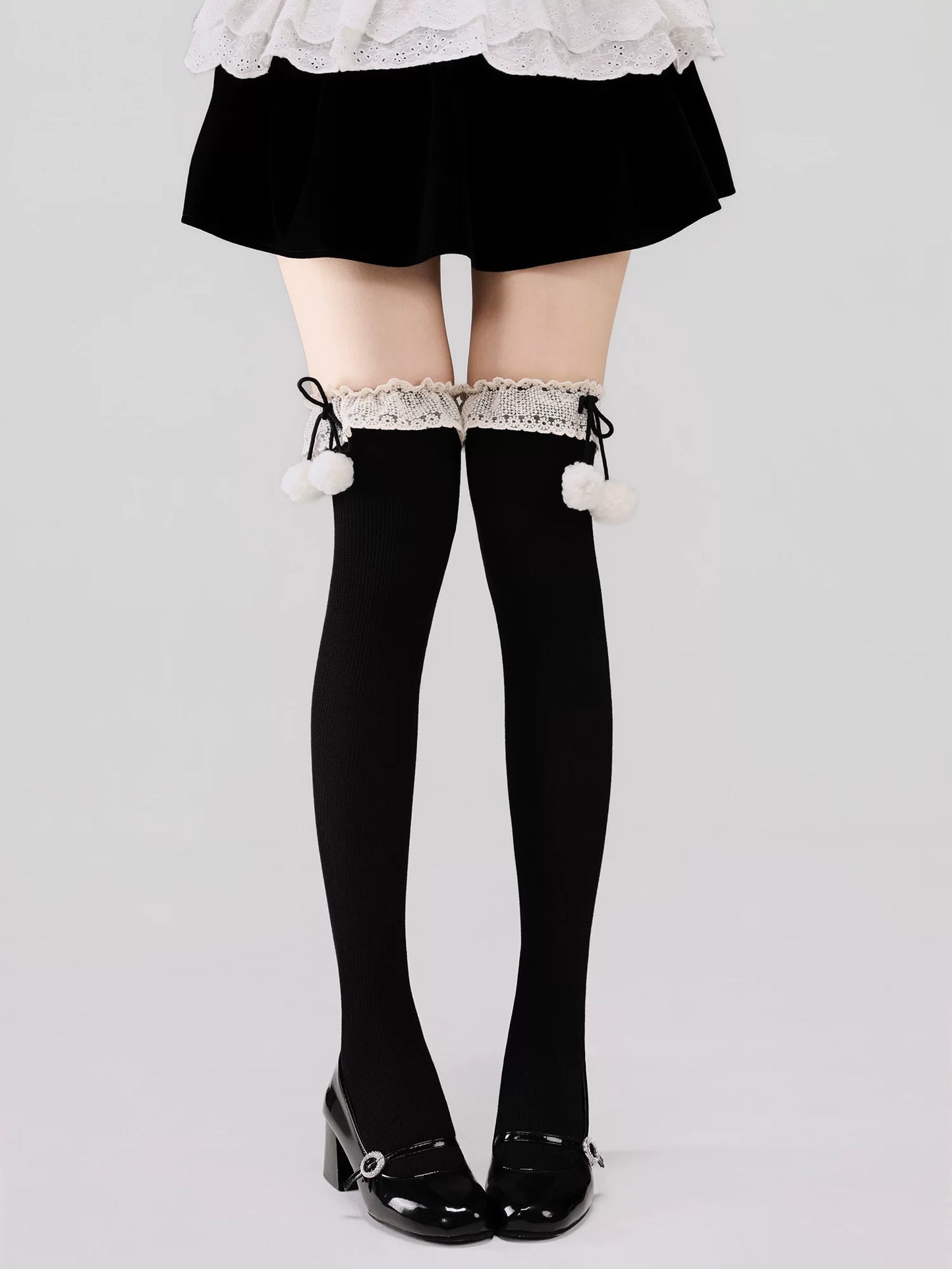 Black thigh-high stockings with white pom-poms and lace trim worn with a black skirt and shoes on a light gray background.