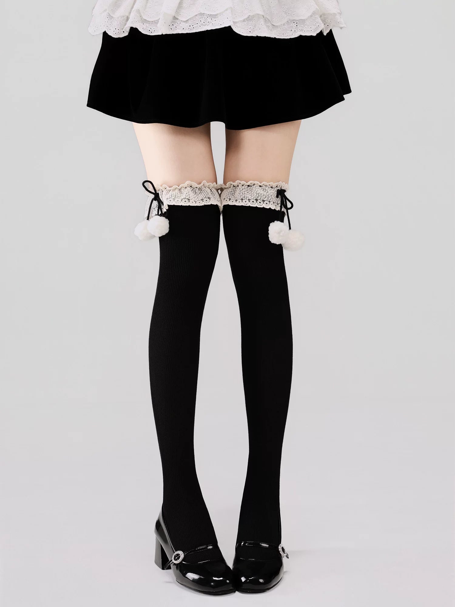 Black thigh-high stockings with white pom-poms and lace trim worn with a black skirt and shoes on a light gray background.