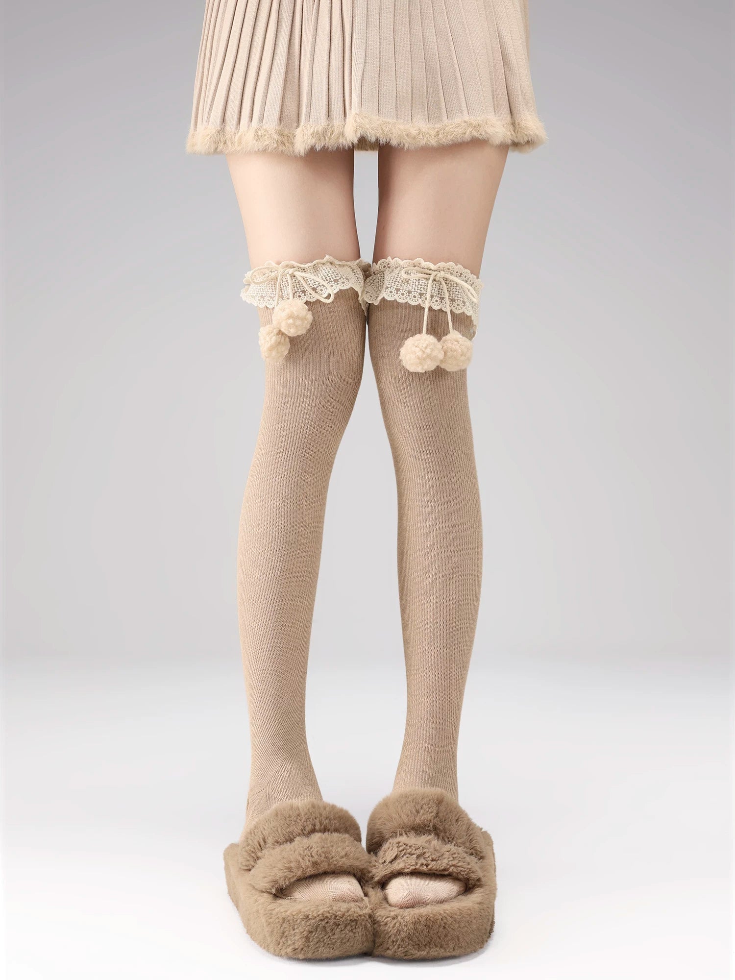 Person wearing beige tights with lace details and fluffy pom-poms, paired with a skirt and slippers on a plain background.