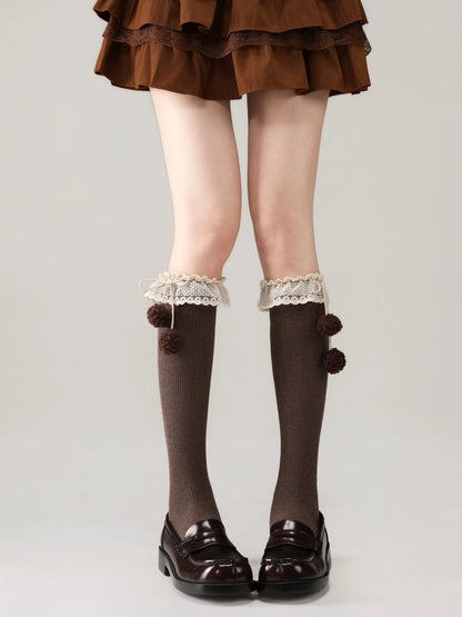 Brown knee-high socks with lace trim and pom-poms worn with a brown skirt and shoes on a light gray background.