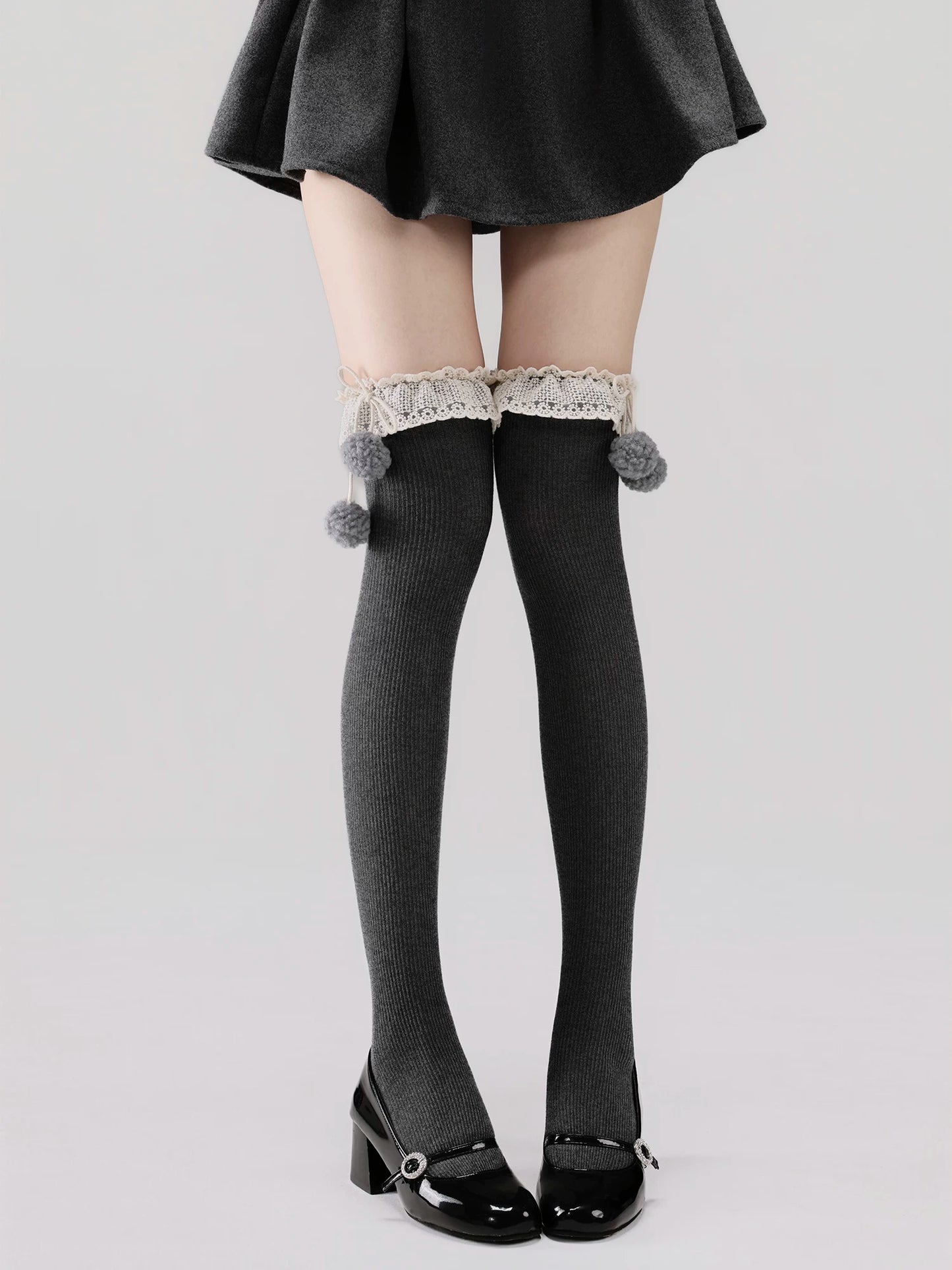 Person wearing black knee-high socks with lace and pom-poms, paired with a black skirt and shoes on a light gray background.