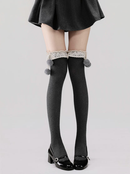 Person wearing black knee-high socks with lace and pom-poms, paired with a black skirt and shoes on a light gray background.