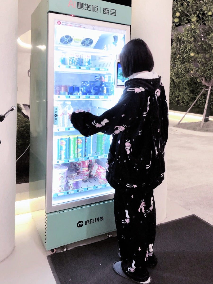 A person in a black and white patterned pajama set stands in front of a mint green beverage vending machine, reaching toward the shelves filled with bottled drinks and snacks.