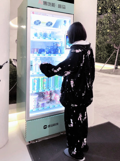 A person in a black and white patterned pajama set stands in front of a mint green beverage vending machine, reaching toward the shelves filled with bottled drinks and snacks.