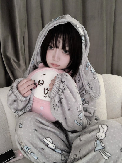 A person wearing a gray bunny-print hooded pajama set sits on a light-colored sofa, holding a pink plush toy with a cartoon face.