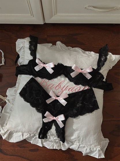 Black lace lingerie set with pink bows on a white pillow, placed on a wooden floor.