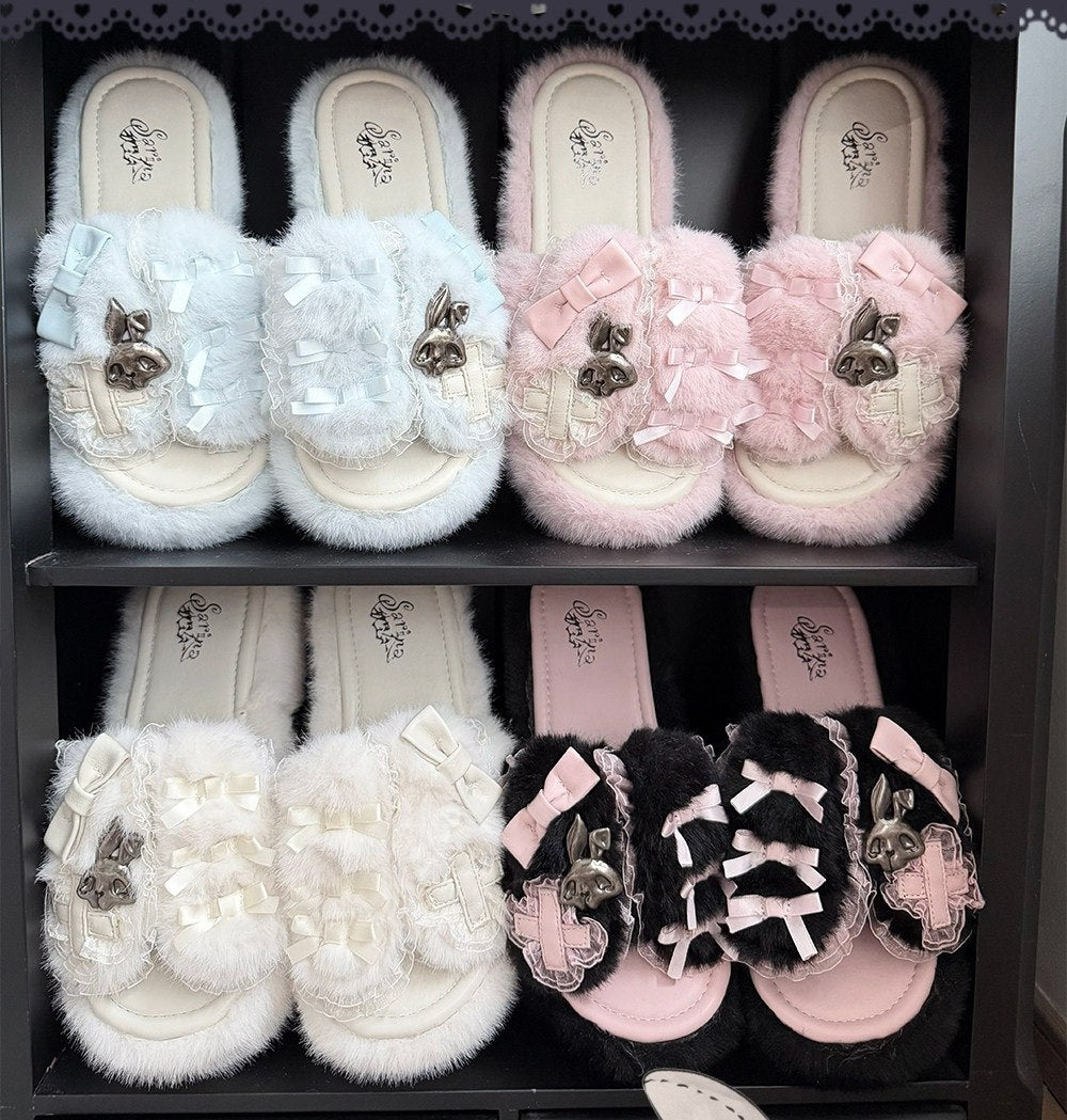 Four pairs of fuzzy slide sandals displayed on dark shelves, each with a large bow and decorative embellishment on the strap. The colors shown are light blue, light pink, off-white/cream, and black with pink and white accents.