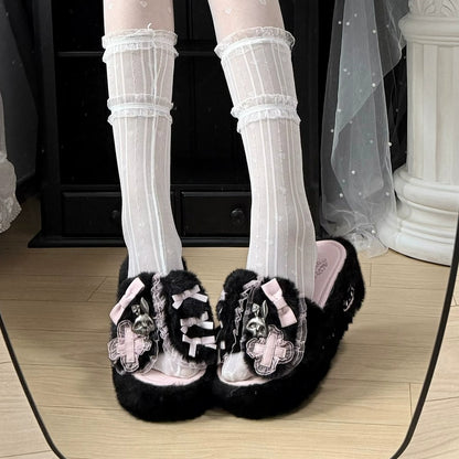 Black fuzzy platform slippers with pink bows, skull details, and lace-up front embellishments, shown on feet with white striped socks in an indoor setting.